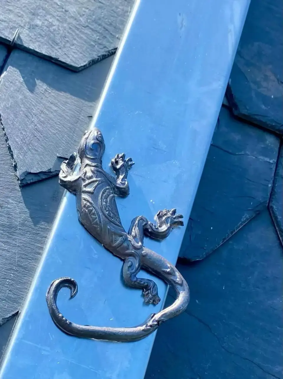 Metalen hagedisornament op een metalen dakstrook tussen donkere leien
